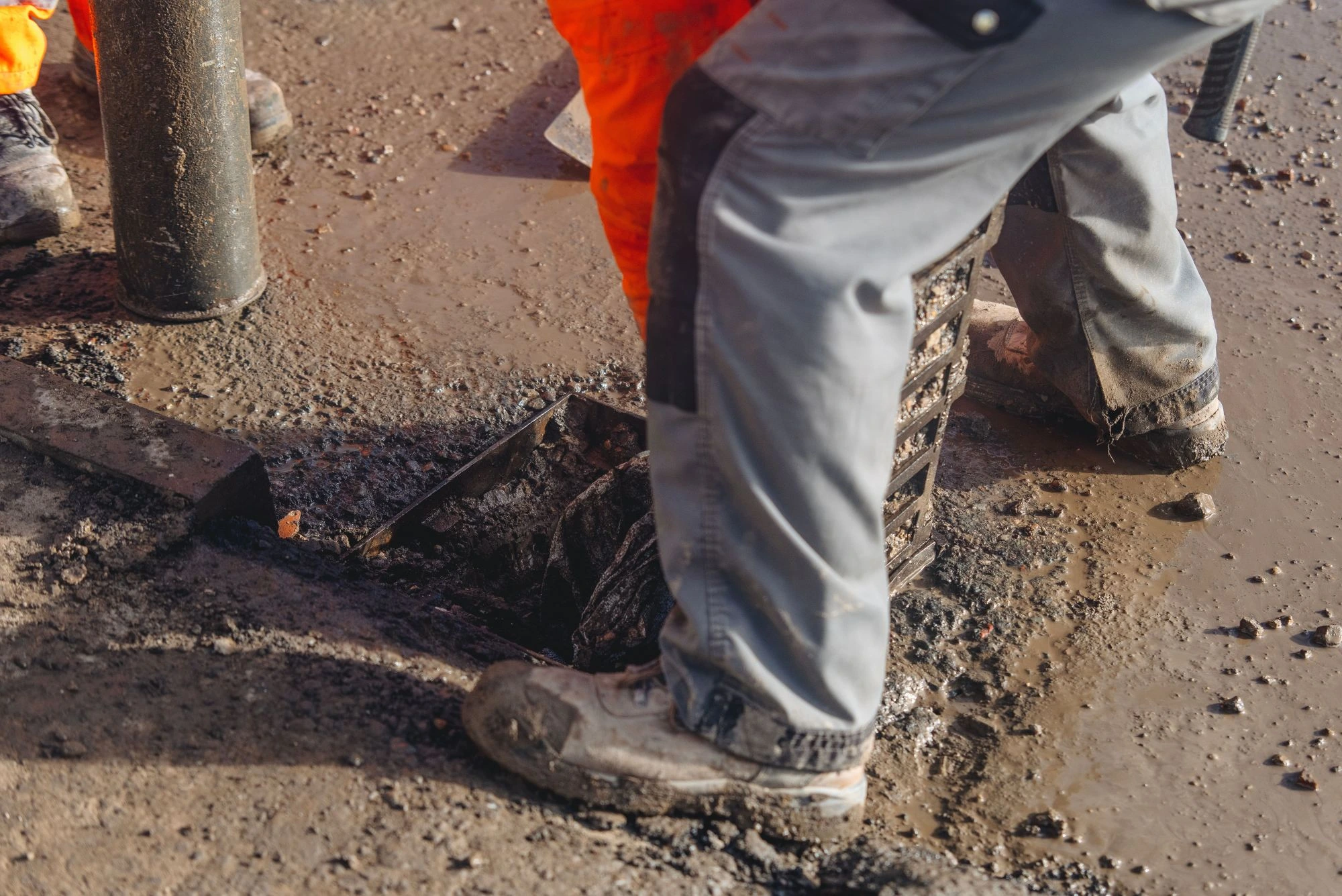 A construction worker in grey and orange work pants standing over an open road gully in a muddy, wet construction area.